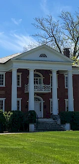 The image shows a classic two-story red brick house with white columns, surrounded by lush greenery and trees under a clear blue sky.